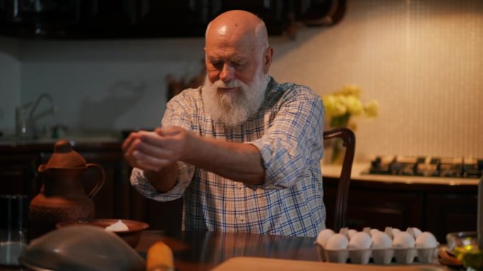 一位头发花白、留着胡子、穿着衬衫的老人，坐在一所现代住宅的厨房里的桌子旁。那人从碗里拿起面粉，倒在桌