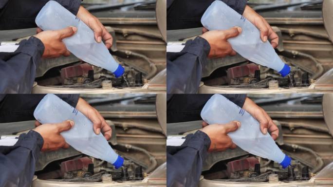 汽车修理工正在为汽车蓄电池机械维修倒去离子纯化蒸馏水。近距离手男子拿去离子蒸馏水瓶在车库汽车移动汽车