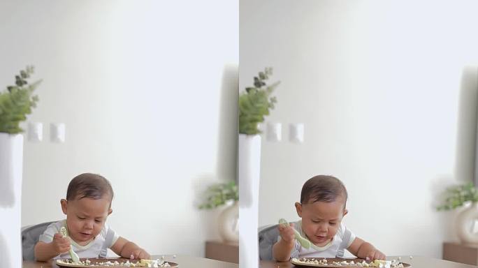 婴儿坐在桌子旁用手拿着预压勺吃东西。