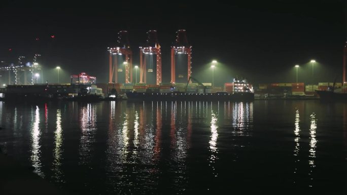 夜晚港口的景象与货物集装箱起重机，灯光反射在水中。月光下集装箱货物作业，吊车活跃。平静的水面映照出正