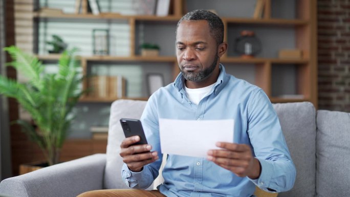 成熟的非洲裔美国人坐在家里的房间里用智能手机支付账单