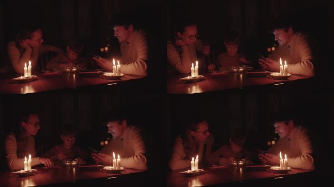 停电时一家人拿着蜡烛坐在家里。孩子在画画，男人在用移动电源给智能手机充电。能源危机概念。