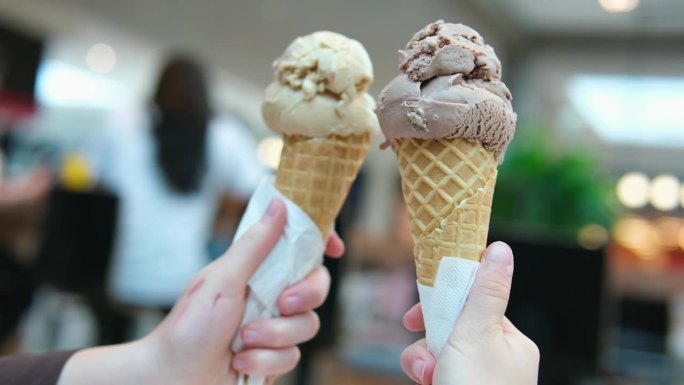 两份冰淇淋甜筒以商场为背景，两个女孩碰杯在一起，拿着棕色的眼镜，游戏交流，友谊长存，吃着美味的甜点。