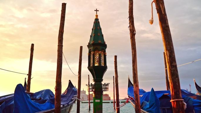 在威尼斯柔和的粉黄色日出中，绿色灯笼教堂的十字架穿过摇曳的贡多拉和灯笼矗立在蔚蓝的泻湖中