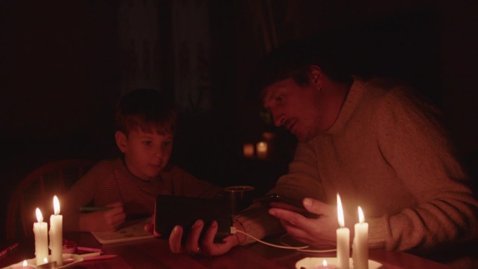 停电时一家人拿着蜡烛坐在家里。孩子在画画，男人在用移动电源给智能手机充电。能源危机概念。