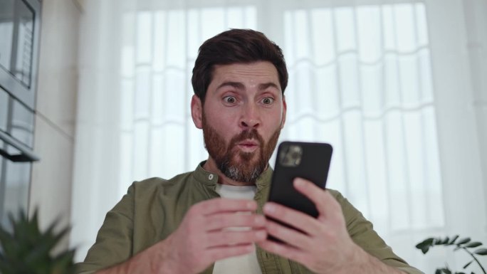 在现代智能手机上发布好消息时，大胡子男人睁大眼睛，张大嘴巴。穿着休闲服的白种男性自由职业者，在家里远