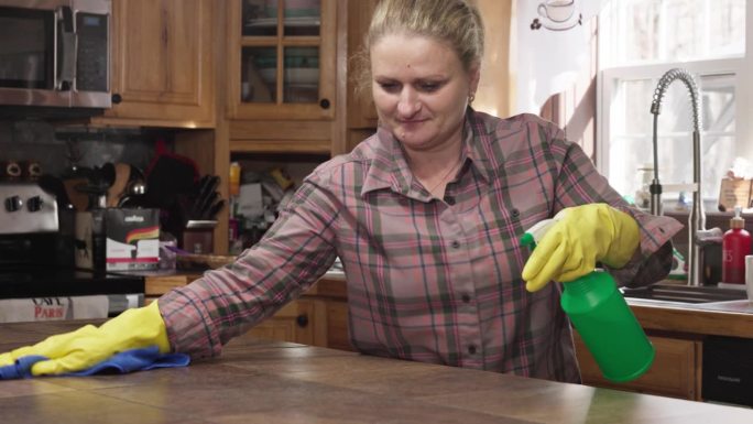 成熟的金发女环卫工人用喷雾瓶擦拭厨房桌子。手持摄像机运动