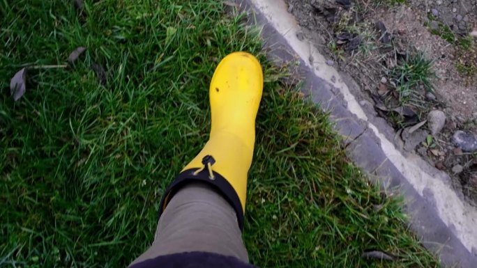 园丁脚踩黄色胶靴，走在绿草地和碎石地上。秋天的季节。特写镜头。从上面看。身体的部分。农夫在农场里走来