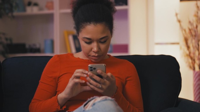 年轻的非裔美国女性坐在家里的沙发上玩手机
