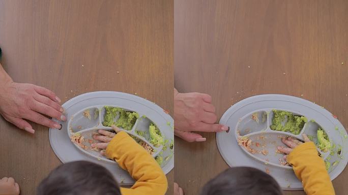 婴儿主导的断奶概念。俯视图，一个婴儿的手从放在木桌上的硅胶盘子里吃东西