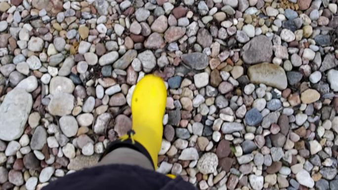 园丁脚踩黄色胶靴，走在碎石地上、道路上。石头表面。秋天的季节。特写镜头。从上面看。身体的部分。农夫在