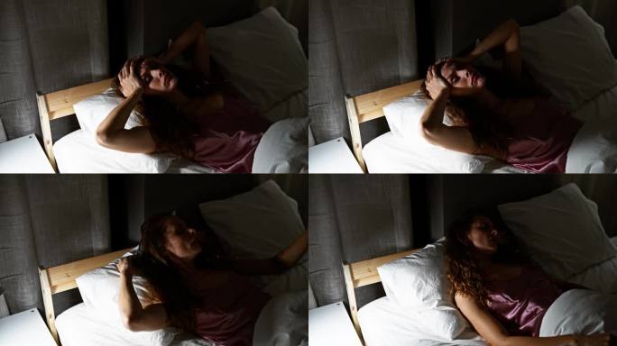 在黑暗的卧室里，一个白人女人躺在床上，捂着自己的头，表现了情感上的混乱。