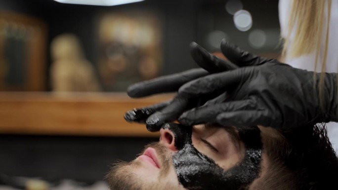 理发师在沙龙里给男顾客敷黑炭面膜做皮肤护理。美容师在男士脸上进行清洁面部护理。美容课程，男士化妆品，