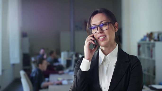 成功的女商人在办公室的肖像，打重要的电话，并与投资者达成协议。在后台使用电脑的公司员工。团队经理使用