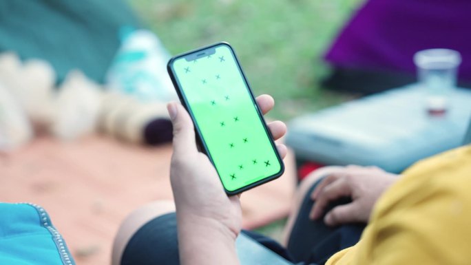 一名男子拿着智能手机，在绿色屏幕的人像模式下，在露营区的帐篷旁边