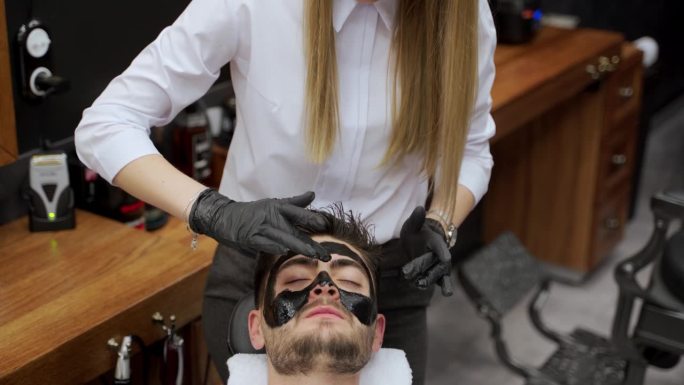 理发师在美容过程中为客户使用黑色面膜清洁毛孔。理发店里时髦的男性护肤程序。女美容师在男士脸上擦去脱皮