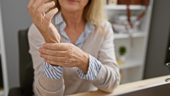 成熟的金发女子在办公室经历手腕疼痛，暗示腕管综合症或类似的重复性劳损。