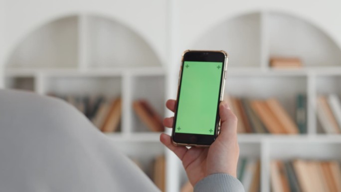 一只手拿着绿色屏幕的智能手机的特写，背面，背景是白色的书架，书架上有很多书