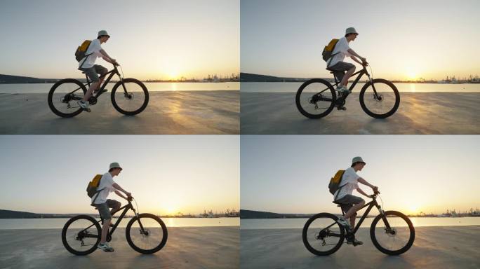 夏天炎热的日落时分，在城市港口码头的海边，少年戴着背包帽骑着自行车的剪影。一代的生活方式。自由。运动