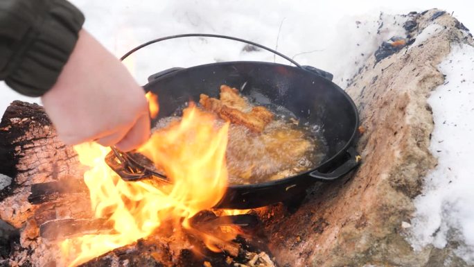 冬雪中，男子在明火上用木铲在金属锅里翻炒炸鸡翅。在野外烹饪，在寒冷中与大自然独处。