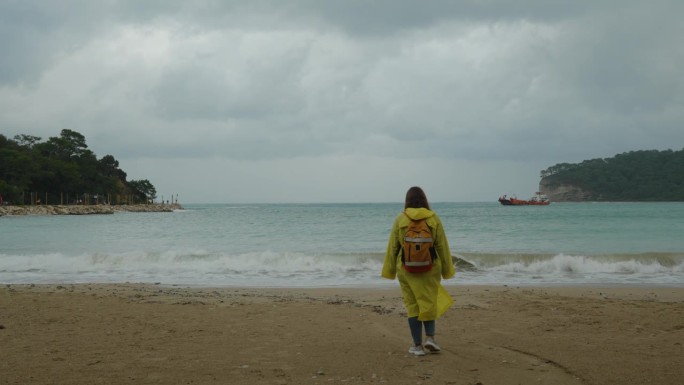 岩石间的海湾，远处的一艘船，一个穿着黄色雨衣的女人迎着风沿着沙滩走。多云的天气。