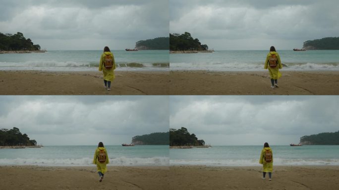 岩石间的海湾，远处的一艘船，一个穿着黄色雨衣的女人迎着风沿着沙滩走。多云的天气。