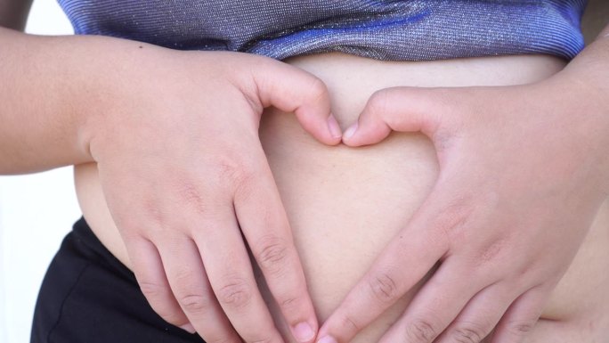 胖女人露出心形的手放在肚子上，为身体护理，怀孕饮食。亚洲年轻女性在暴饮暴食中存在肥胖问题。4 k