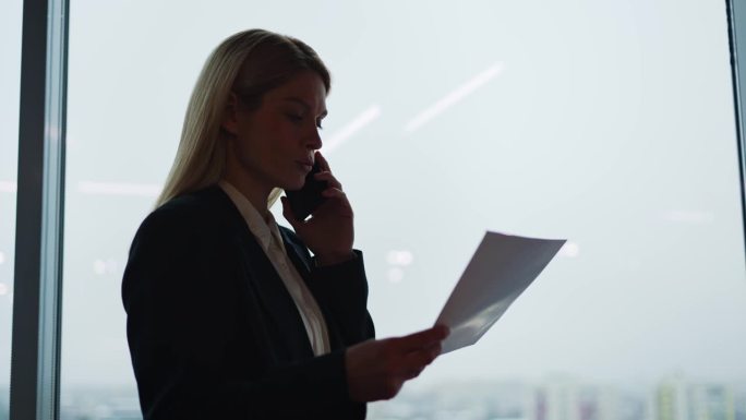 一个女商人的剪影在一个现代化的玻璃办公室里用手机交谈，背景是摩天大楼的窗户。成功自信的女商人站在室内