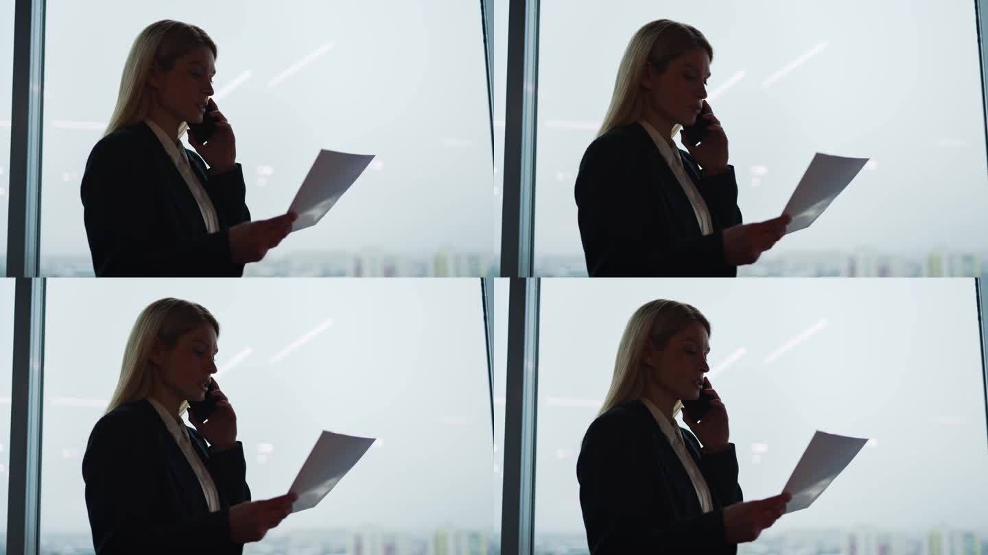一个女商人的剪影在一个现代化的玻璃办公室里用手机交谈，背景是摩天大楼的窗户。成功自信的女商人站在室内