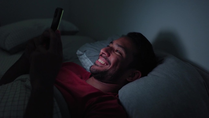 男人，晚上和电话在床上，而开心和笑的搞笑视频，表情包和阅读博客在家里。男人，带着微笑的人和智能手机，