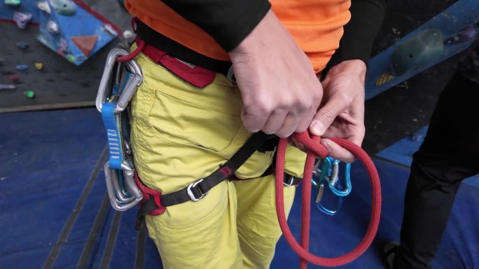 为了保证攀爬的安全开始，攀岩者熟练地运用攀岩结技术在攀岩墙前系好绳子。
