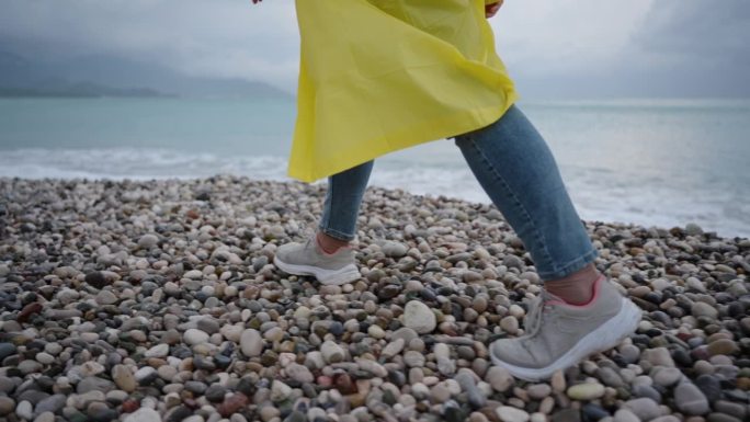 暴风雨中汹涌的大海，不祥的气氛，穿着牛仔裤和亮黄色雨衣的女人的腿，沿着卵石滩散步。慢动作。