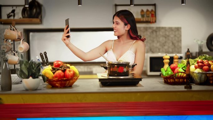 快乐的印度妇女站在现代化的厨房里，拿着木铲搅拌素食汤，在家里的火锅里准备早餐。微笑的女性使用智能手机