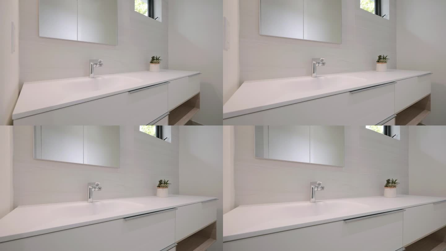 现代简约的浴室水槽在光滑的白色台面。小的盆栽植物为空间增添了绿色，上面的镜子增加了房间的亮度