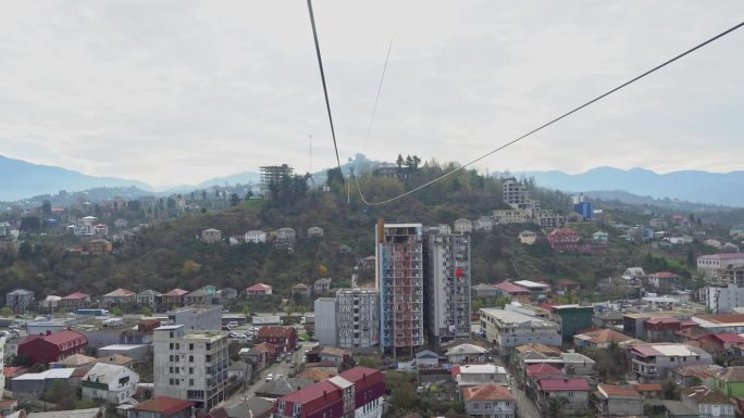 从索道的船舱里，沿着缆索上升，俯瞰城市，背景是群山。悬挂架沿滚子接头移动。山地缆车