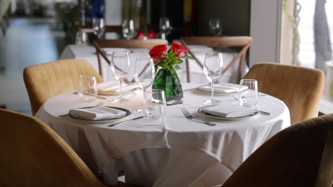 餐厅里摆放整齐的圆桌，铺着白色桌布。每个地方包括盘子、餐巾纸和两个杯子。绿色花瓶里的一束红玫瑰