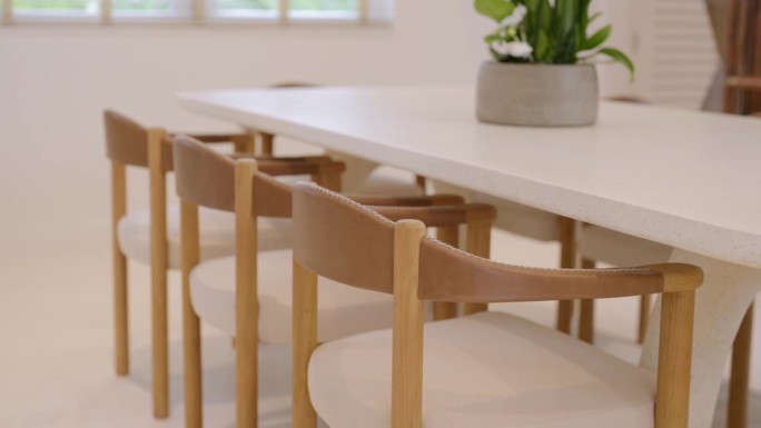 简约的现代用餐区，白色长桌周围是米色靠垫的木椅。郁郁葱葱的绿叶盆栽植物放在桌子上的灰色花盆