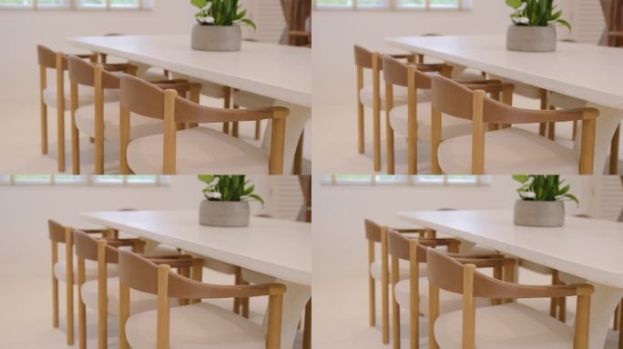 简约的现代用餐区，白色长桌周围是米色靠垫的木椅。郁郁葱葱的绿叶盆栽植物放在桌子上的灰色花盆