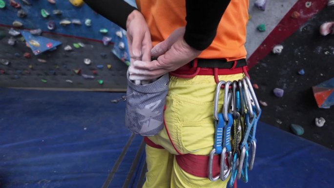 穿着黄色裤子的男攀岩者在攀岩墙前把手伸进镁粉包里准备攀岩。安全设备挂在攀登吊带上。