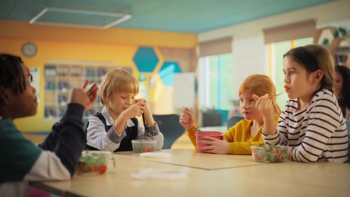 小学生在午休时间吃健康食物。聪明多样的孩子们在现代色彩缤纷的教室里享受乐趣，交谈和美味的沙拉餐