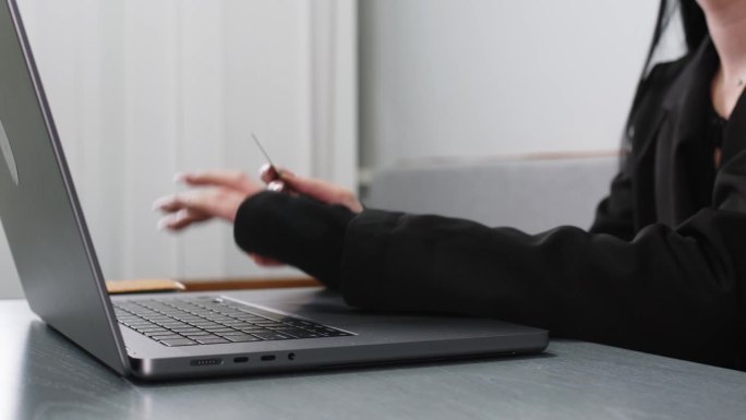 一名年轻女子手持银行卡，在房间的桌子上通过笔记本电脑进行网上购物的特写。网上购物和使用信用卡的概念。
