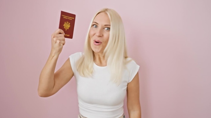 震惊的年轻金发女子，难以置信地张大嘴巴，在孤立的粉红色背景上抓着法国护照，情绪失控，害怕但又惊讶。