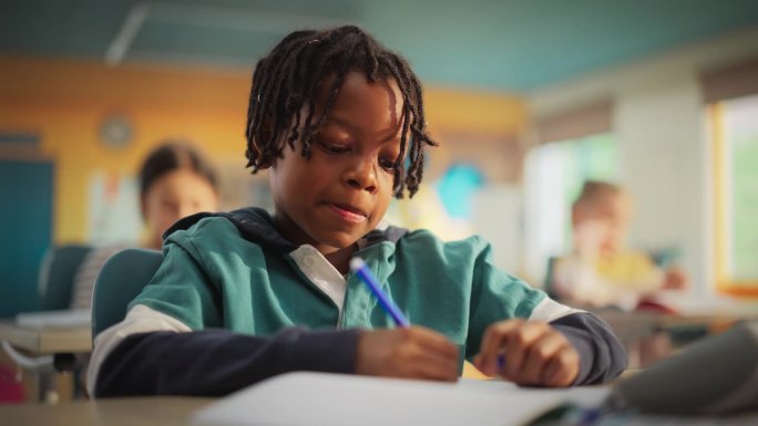 一个可爱的黑人小男孩的肖像，带着可爱的微笑坐在课桌后面。年轻的学生专注于一堂课，和其他孩子一起听老师