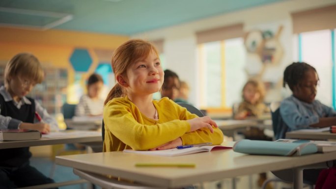 一个可爱的姜黄色头发的女孩坐在教室的桌子后面。小学生专心听讲，和其他孩子一起听老师讲课