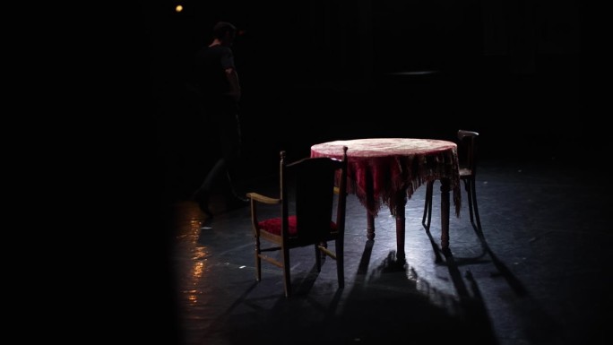 演员走过舞台，旁边是一张旧桌子和椅子。