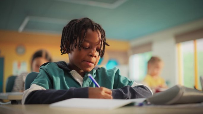 聪明的留着脏辫的非洲小男孩坐在小学的桌子后面。细心的非洲年轻人在笔记本上写下笔记，找到解决练习的方法