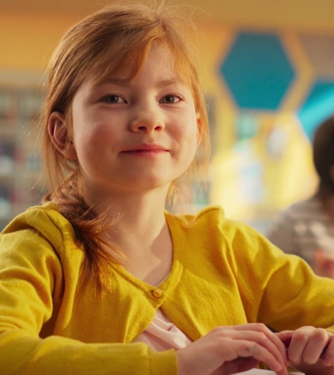 竖屏:一个可爱的红发女孩坐在课桌后面的肖像。小学生微笑着看着镜头。在现代小学接受教育的孩子