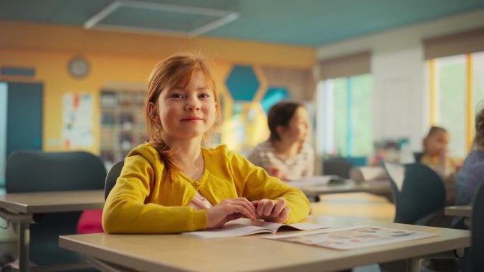 一个可爱的红发女孩坐在课桌后面的肖像。小学生微笑着看着镜头。在现代小学接受教育的孩子