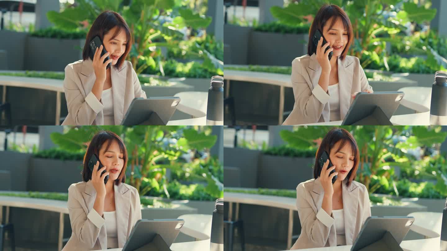 年轻的亚洲女商人一边用平板电脑工作一边打电话，她坐在户外，展示了高效的多任务处理。户外可持续经营。