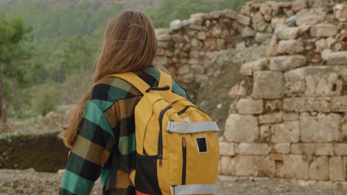一个孤独的女旅行者背着背包，在一座古老的建筑废墟的背景下行走，背景是一片森林。后面的观点。
